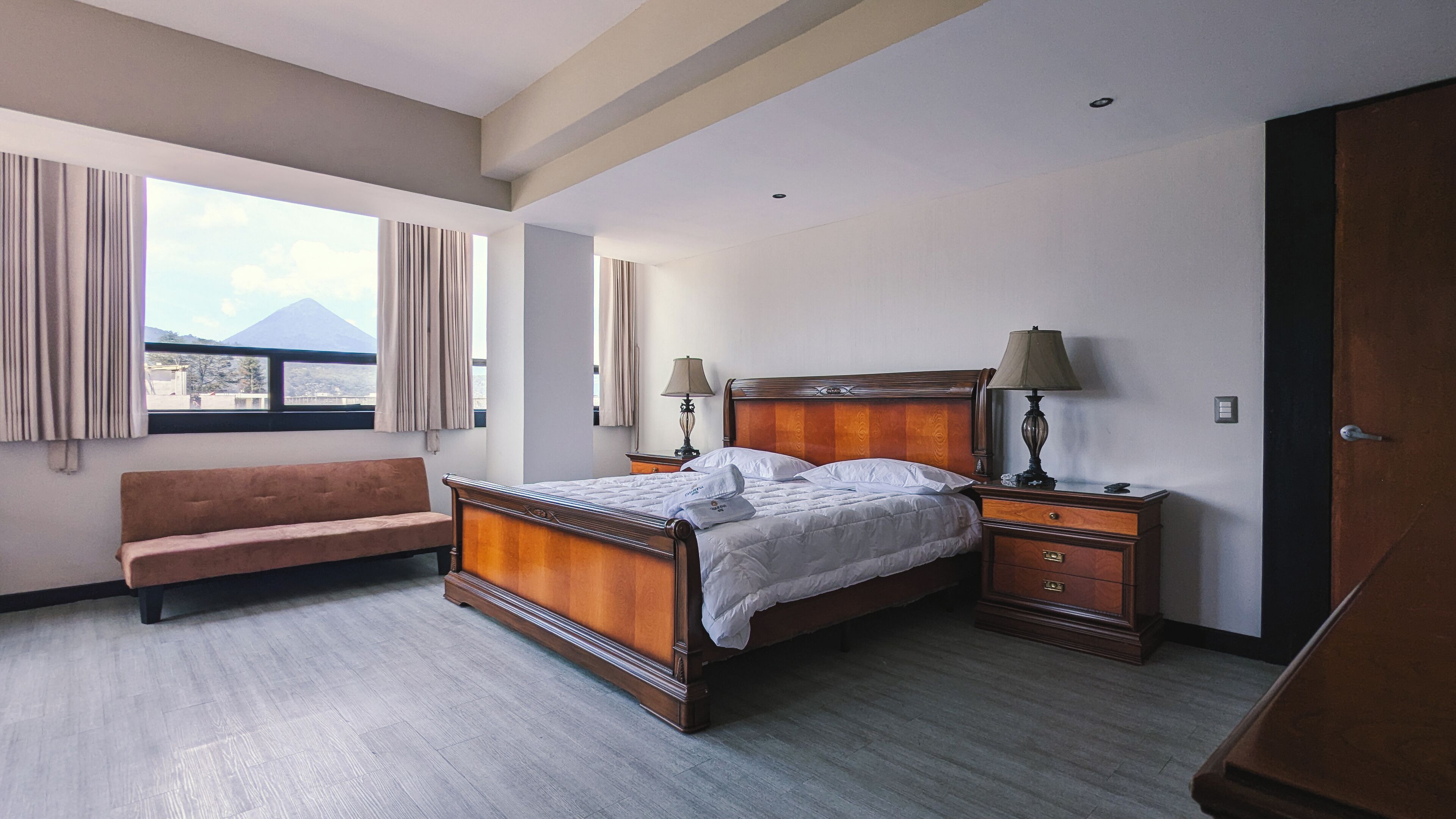 A hotel room with a wooden bed, two bedside tables with lamps, a bench, and a window with curtains.