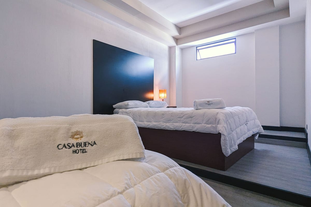 A hotel room with two beds, a large dark artwork on the wall, a window, and a nightstand with a lamp.