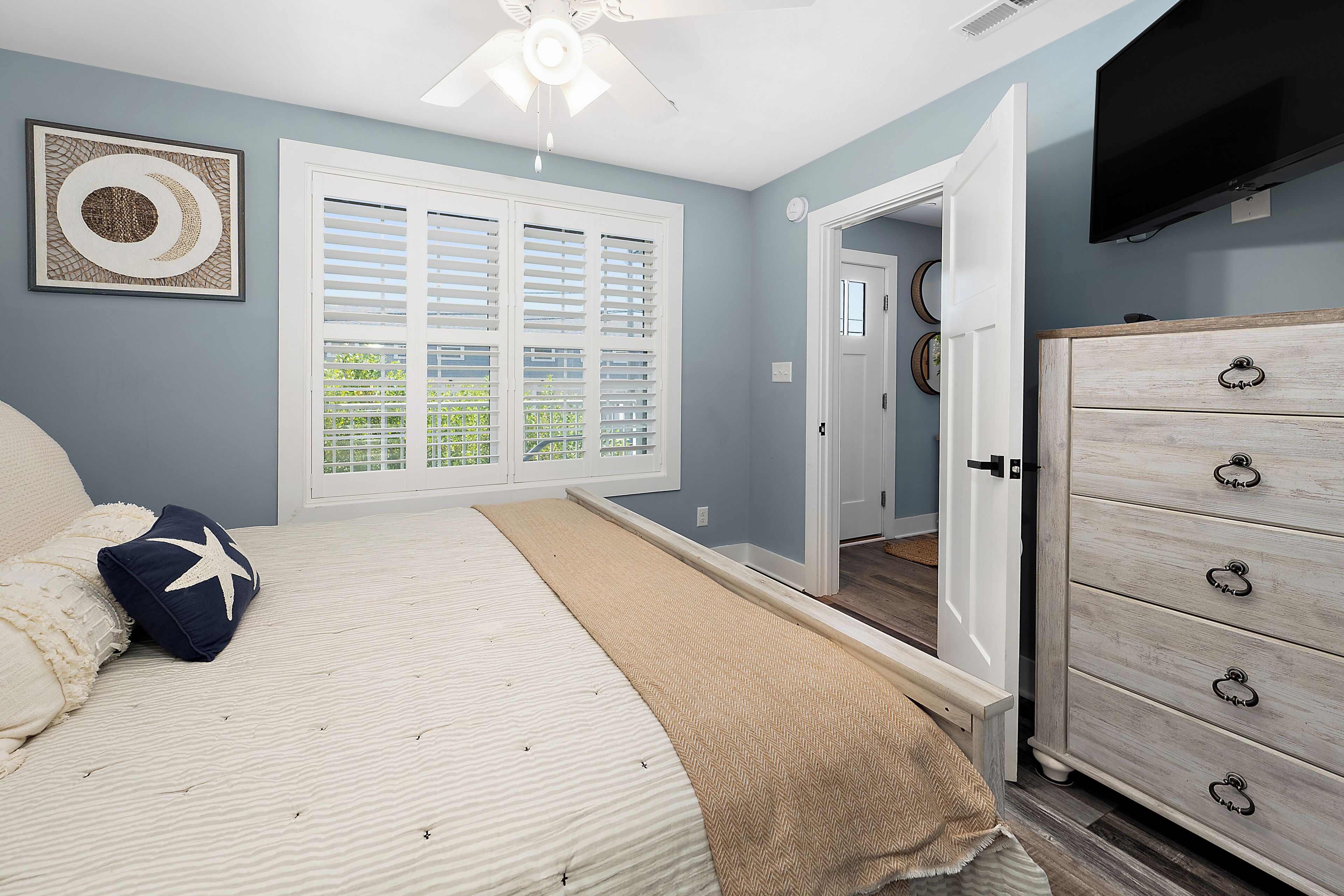 A bedroom with a bed, a dresser, a ceiling fan, a window with blinds, and a door leading to another room.