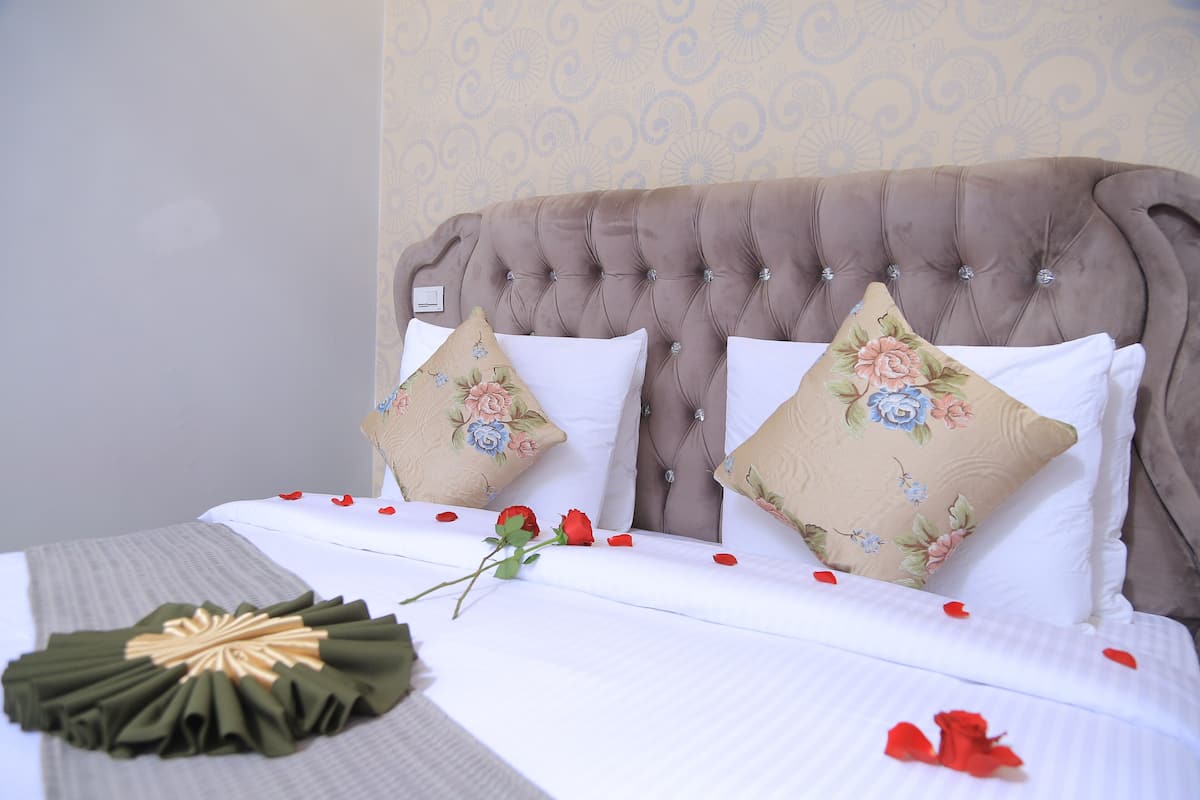 A neatly made bed with floral pillows, a bouquet of green flowers, and scattered rose petals.