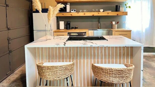 A kitchen with a central island, two woven chairs, and wooden shelves above the counter. - Guest House | King Bed | Zen Retreat (Irving)
