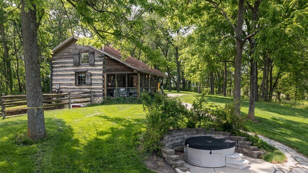 A rustic log cabin with a porch, surrounded by lush greenery and a stone fire pit. - Green Plains Cabin (South Charleston)
