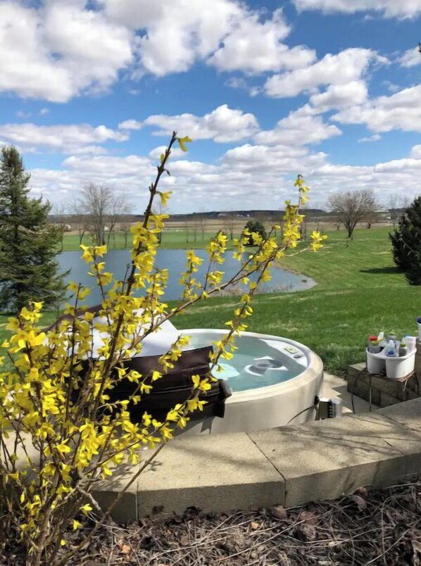 A hot tub with a circular design, surrounded by a concrete deck, with a view of a lake and a grassy area in the background. - Green Plains Cabin - circa 1855 (South Charleston)