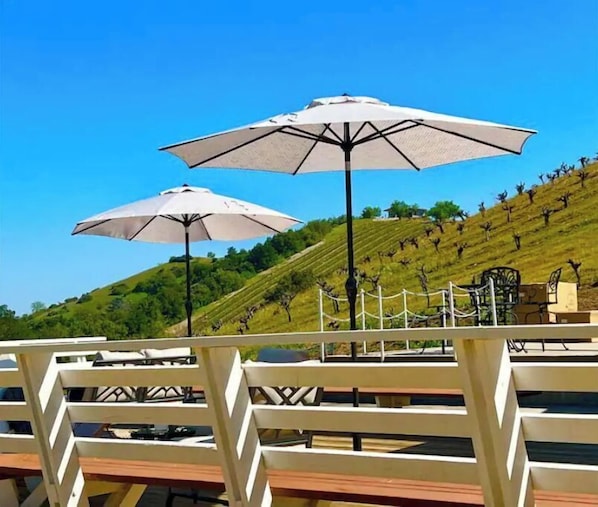 A terrace with two white umbrellas, a wooden fence, and a view of a hill with sculptures.
