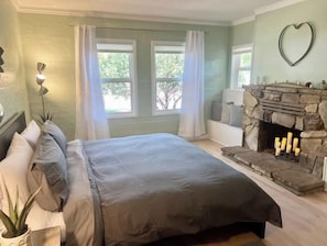 A bedroom with a bed, a stone fireplace, and a view of trees through the windows.