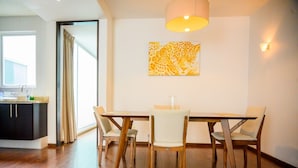 A dining area with a wooden table and chairs, a painting on the wall, and a kitchen in the background.