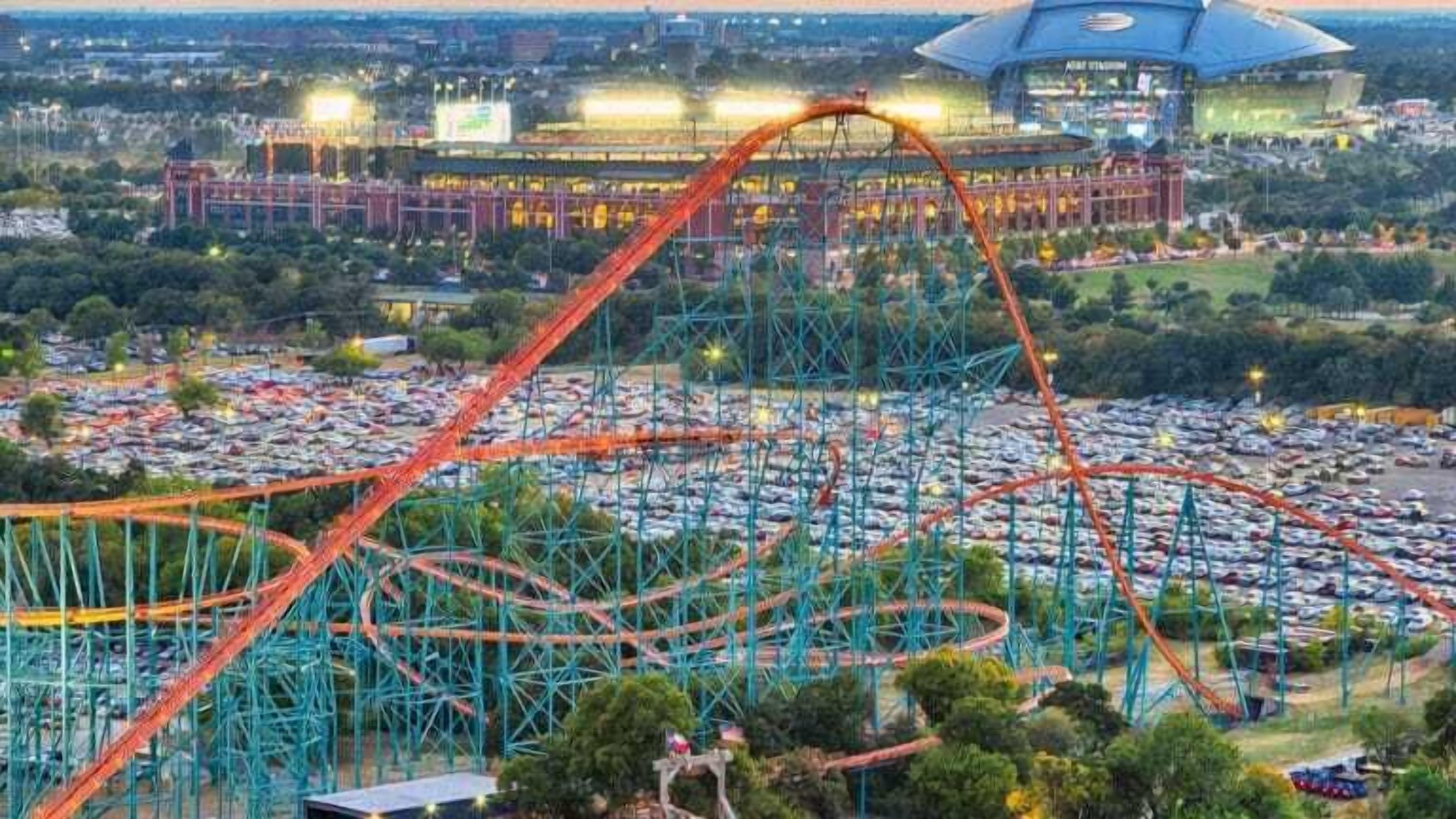 A roller coaster with a steep drop and multiple loops, surrounded by a parking lot and a stadium in the background.