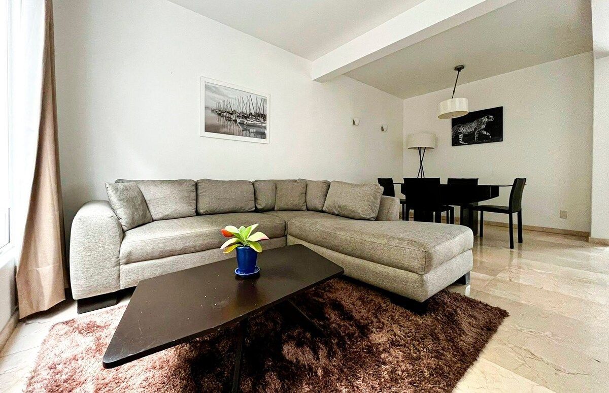 Een moderne woonkamer met een hoekbank, een salontafel, een eettafel met stoelen en kunstwerken aan de muur.