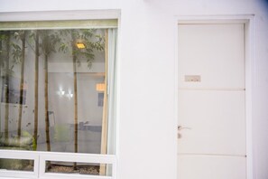 Una habitación con un ventanal grande, una puerta con placa de número y un estante pequeño.