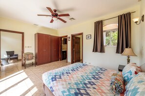 A bedroom with a bed, a ceiling fan, a window with curtains, a bedside lamp, and a view of the bathroom.