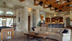 A spacious living area with a pool table, a large sofa, and a coffee table. The room features exposed wooden beams, stone walls, and a view of the kitchen through an open doorway. - Lake Travis Waterfront, Pool, Pool Table, Shuffleboard, Floating Boat Dock (Marble Falls)