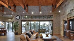 A spacious living area with a large sofa, a coffee table, and a dining table. There is a large window providing a view of the outdoors. - Lake Travis Waterfront, Pool, Pool Table, Shuffleboard, Floating Boat Dock (Marble Falls)
