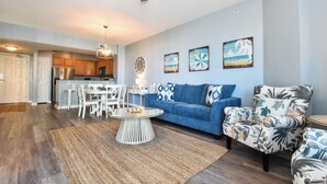 A spacious living room with a blue sofa, a patterned armchair, a dining area with white chairs, and a kitchen in the background. - Beachy Condo w/ Lagoon-Style Pool Views at The Palms of Destin! 💚☀🌴 (Destin)