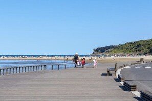 Une promenade en bois qui mène à une plage où l’on voit des gens se promener, avec une falaise rocheuse en arrière-plan.