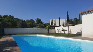A swimming pool with a clear blue water surface, surrounded by a white wall and a grassy area with trees and a palm tree. - Elaia Silvis on the Mediterranean – 14-meter heated pool (TOUROUZELLE 11200)