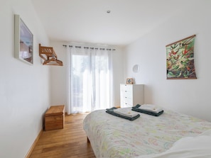 Une chambre avec un plancher en bois, un lit recouvert d’une couvre-lit aux motifs colorés, une commode blanche, un banc en bois, une fenêtre avec des rideaux légers et un tableau accroché au mur.