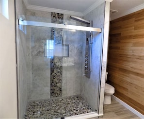 Een douche met glazen wanden, een betegelde vloer, een wandmengkraan en een houten wand naast een toilet.