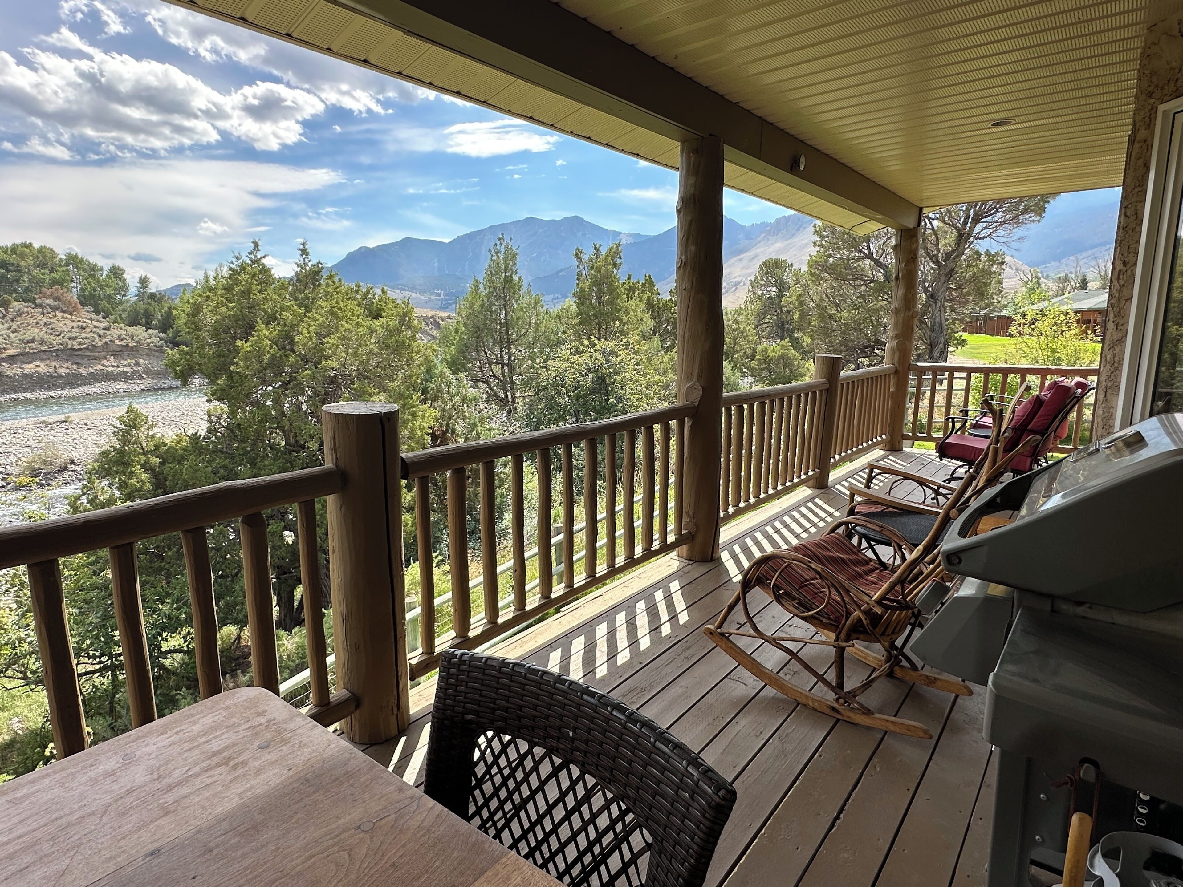 A wooden deck with a view of mountains and a river. There are wicker chairs and a table.