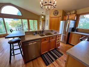 A kitchen with a central island, wooden cabinets, a stainless steel refrigerator, and a large window offering a view of greenery.