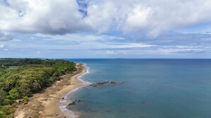 A coastal landscape with a sandy beach, dense greenery, and a clear view of the ocean.