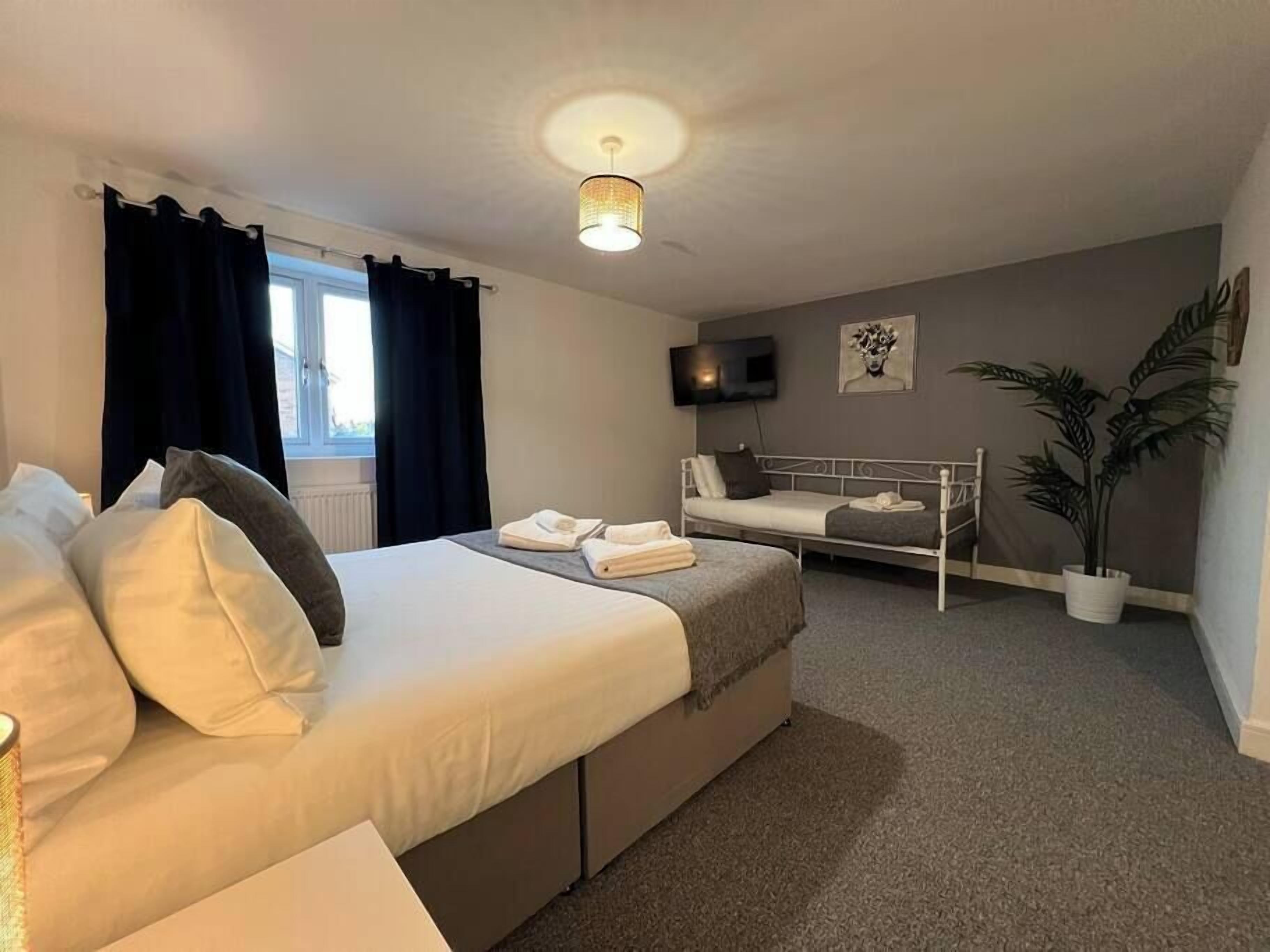 A hotel room with a large bed, a smaller bed, a TV, a plant, and a window with curtains.