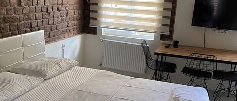 A bedroom with a brick wall, a bed with white linens, a window with blinds, a desk with a TV, and a radiator.