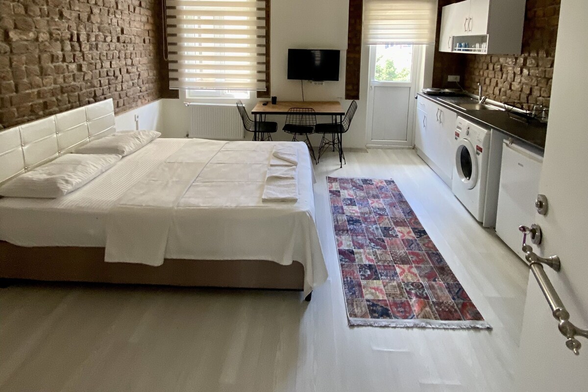 A bedroom with a large bed, a desk, a TV, a washing machine, and a brick wall.