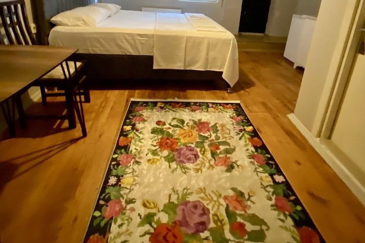 A bedroom with a bed, a wooden floor, a floral rug, a table with chairs, and a ceiling light.