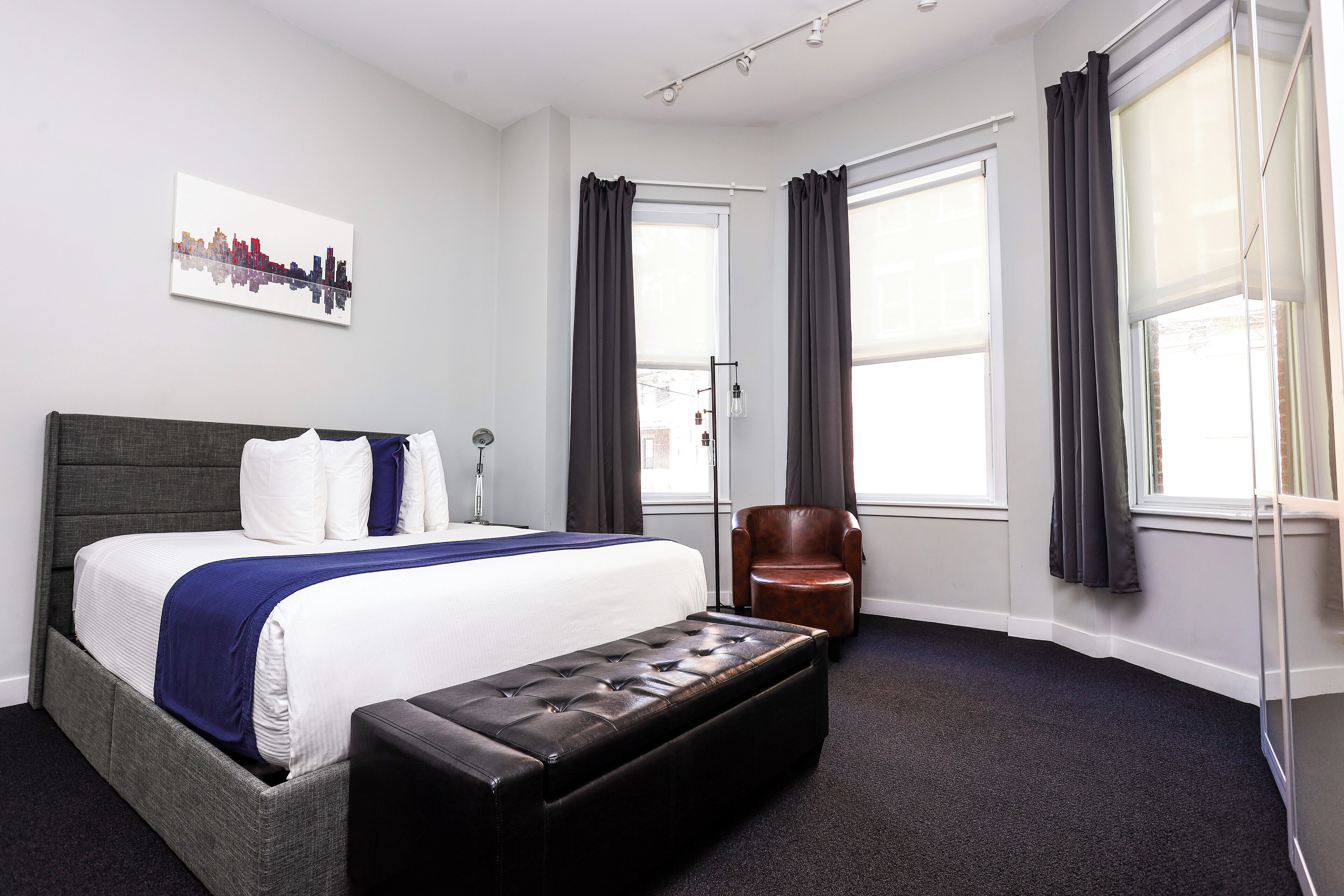 A modern hotel room with a large bed, a bench, a chair, and a cityscape artwork on the wall.