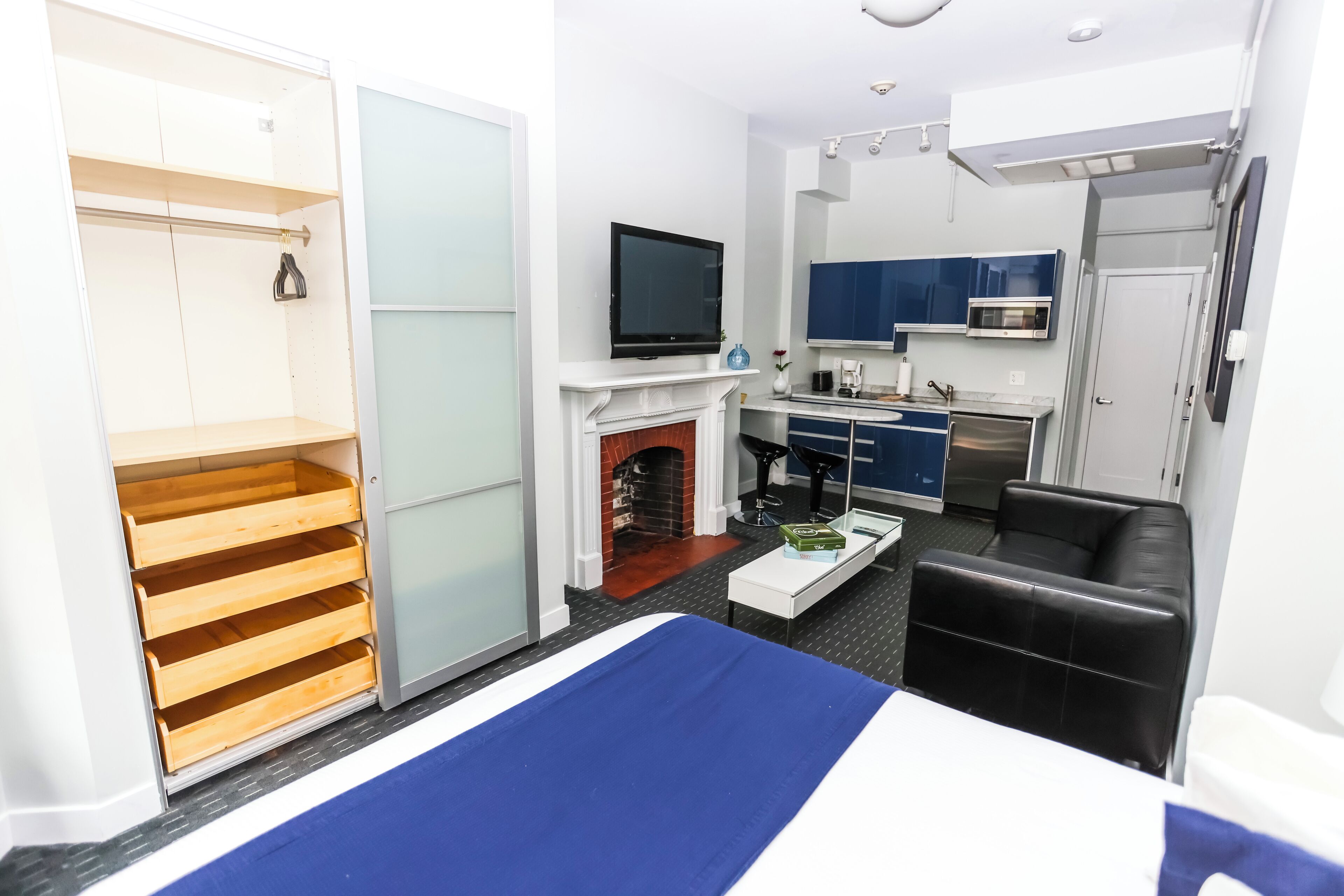 A compact hotel room with a bed, a television, a small kitchen, and a seating area.