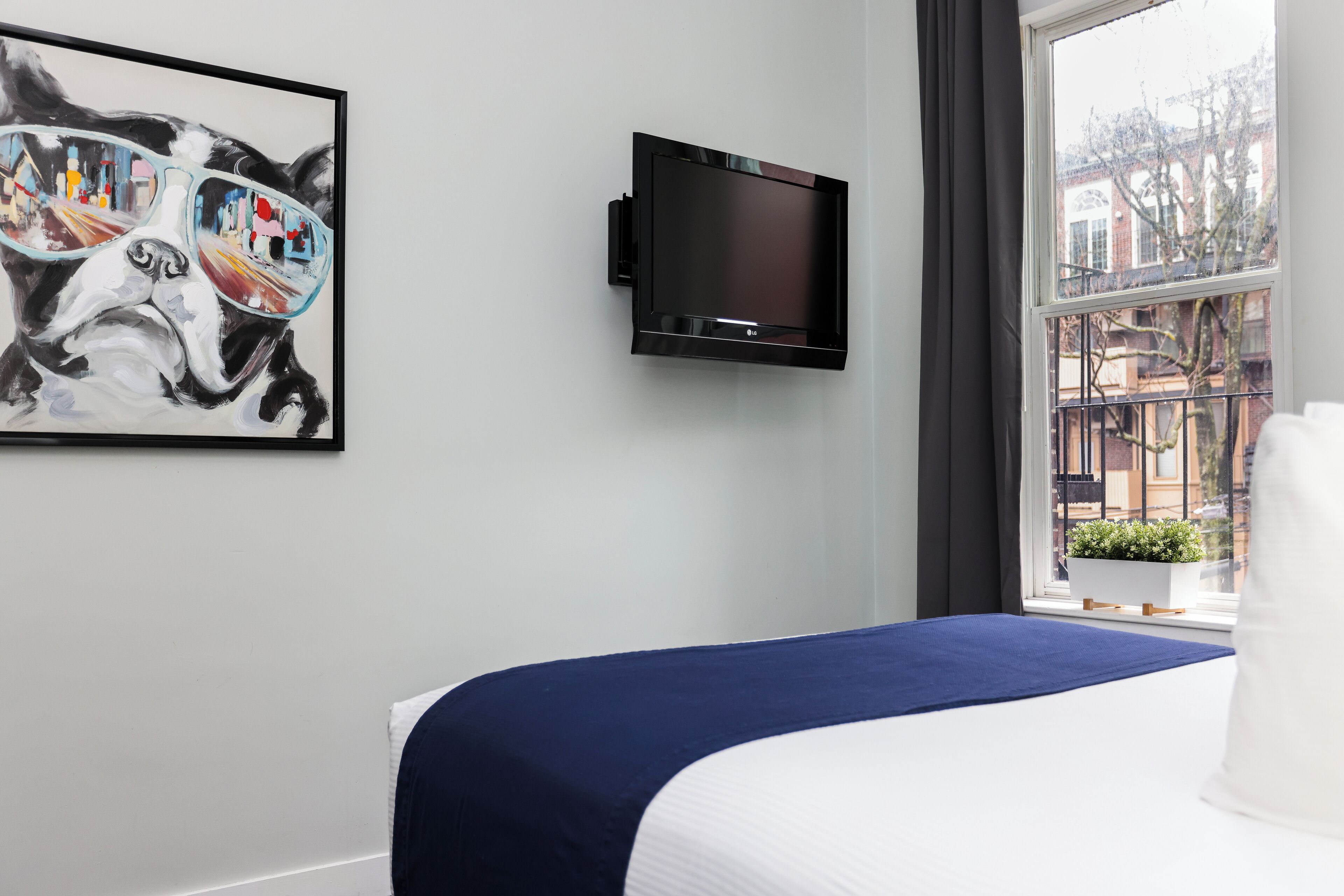 Habitación de hotel con cama, televisor montado en la pared y ventana con vistas a edificios y un balcón.