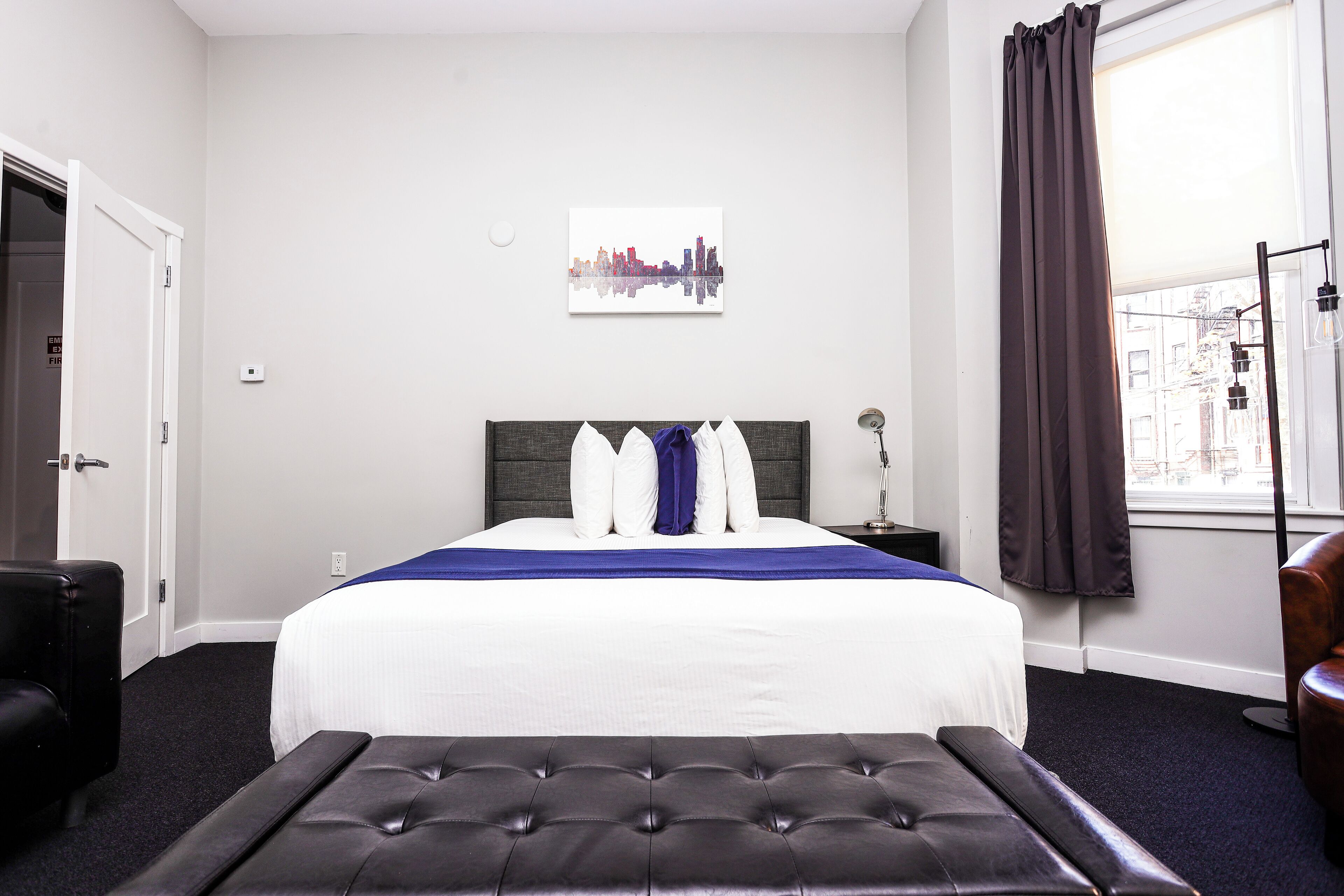 A hotel room with a bed, a bench, a nightstand, a lamp, and a window with curtains.
