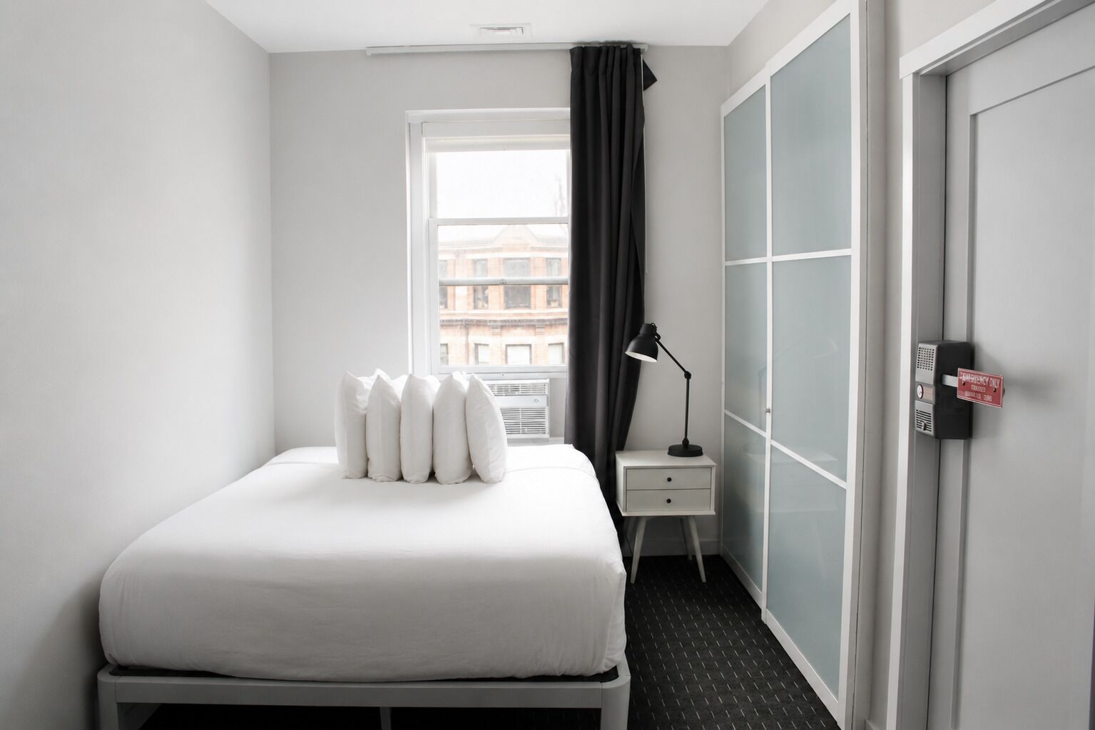A hotel room with a bed, a nightstand with a lamp, a window with curtains, and a door with a sign.