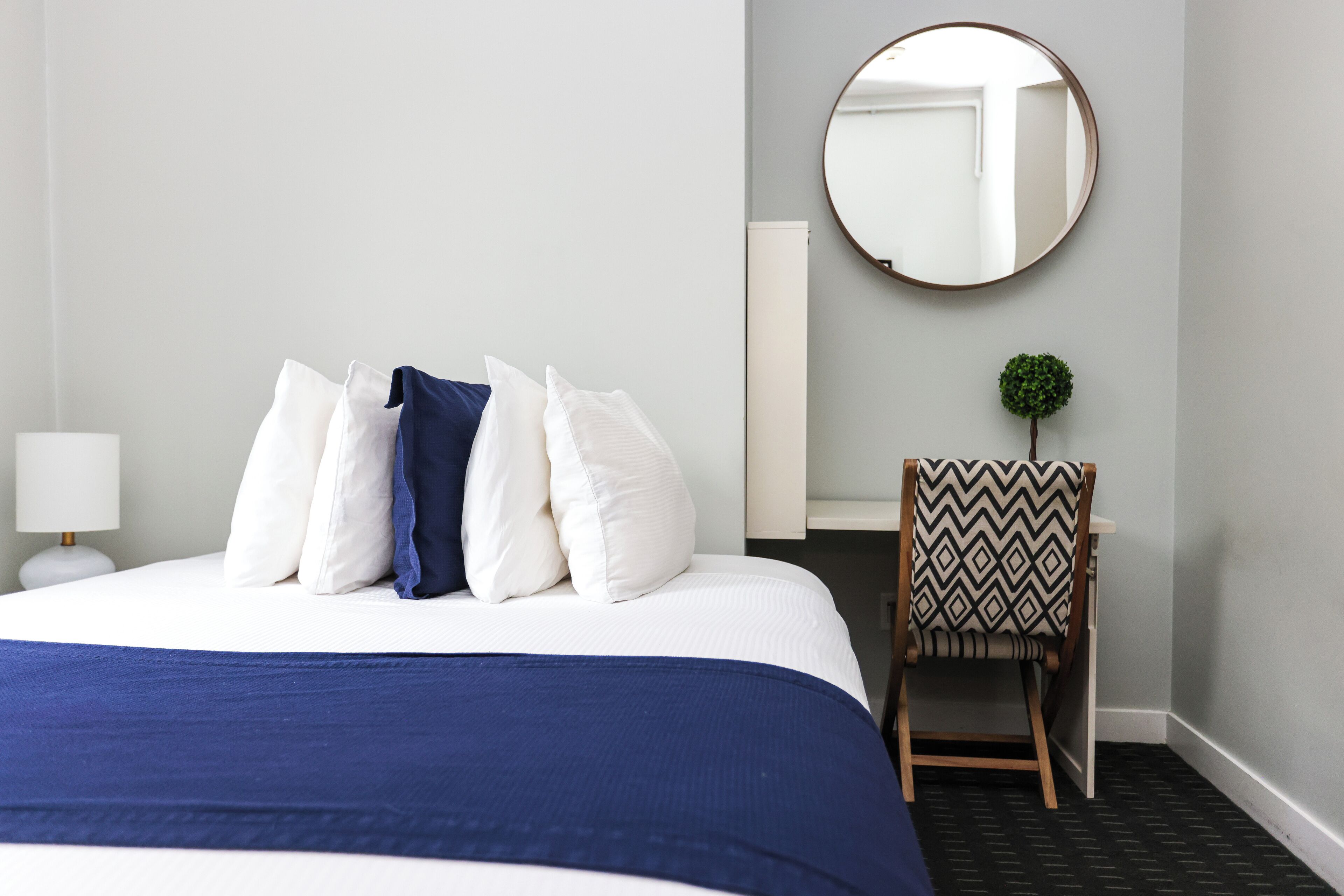 Un lit bien fait, avec une couvre-lit bleue, des oreillers blancs et un petit miroir rond accroché au mur.