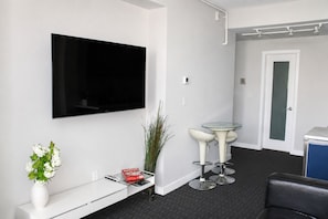 A modern room with a flat-screen TV, a glass table with two chairs, a white console table with a vase and a remote control, and a black leather chair.