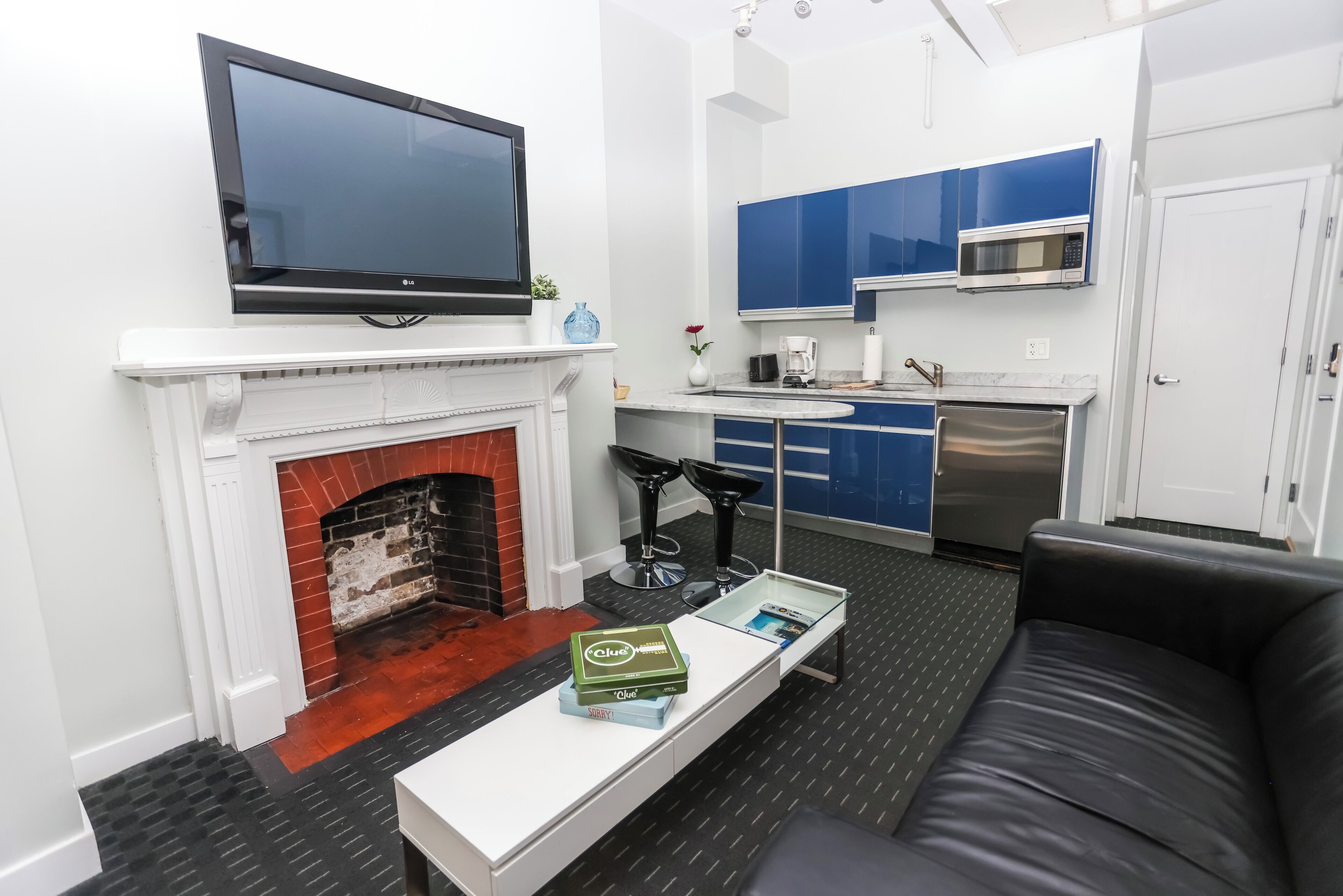 A modern living room with a fireplace, a flat-screen TV, a coffee table with books, a black leather sofa, and a kitchen area with blue cabinets and a microwave.