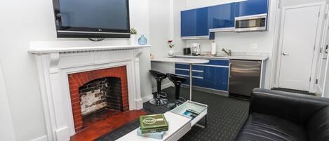 A modern living room with a fireplace, a flat-screen TV, a coffee table with books, a black leather sofa, and a kitchen area with blue cabinets and a microwave.