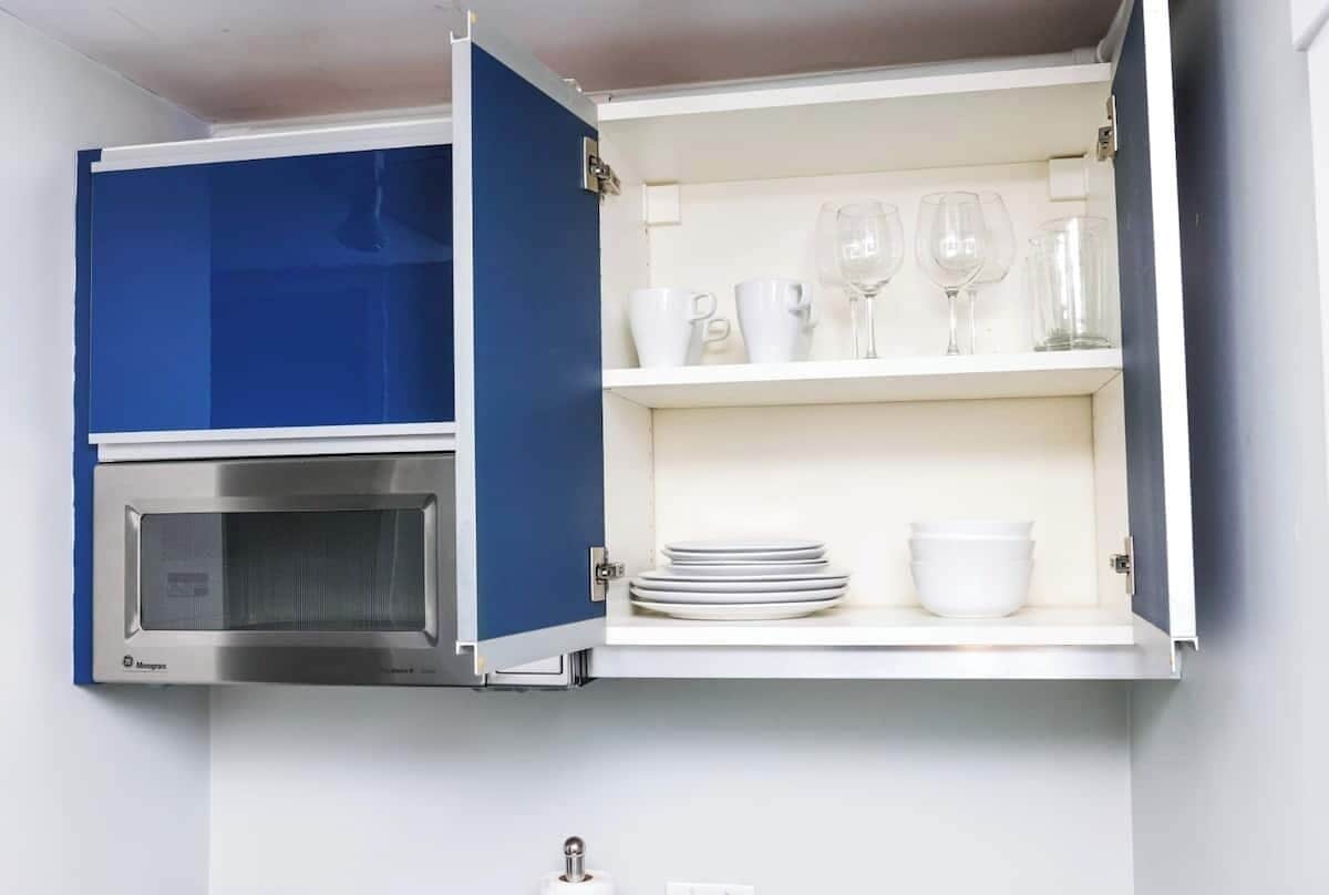 Cocina con microondas incorporado y un armario con estantes que contienen vajilla y vasos blancos.