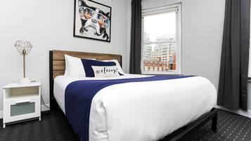 Una camera d'albergo con un letto, un comodino, una lampada, una finestra con tende e vista sulla città.