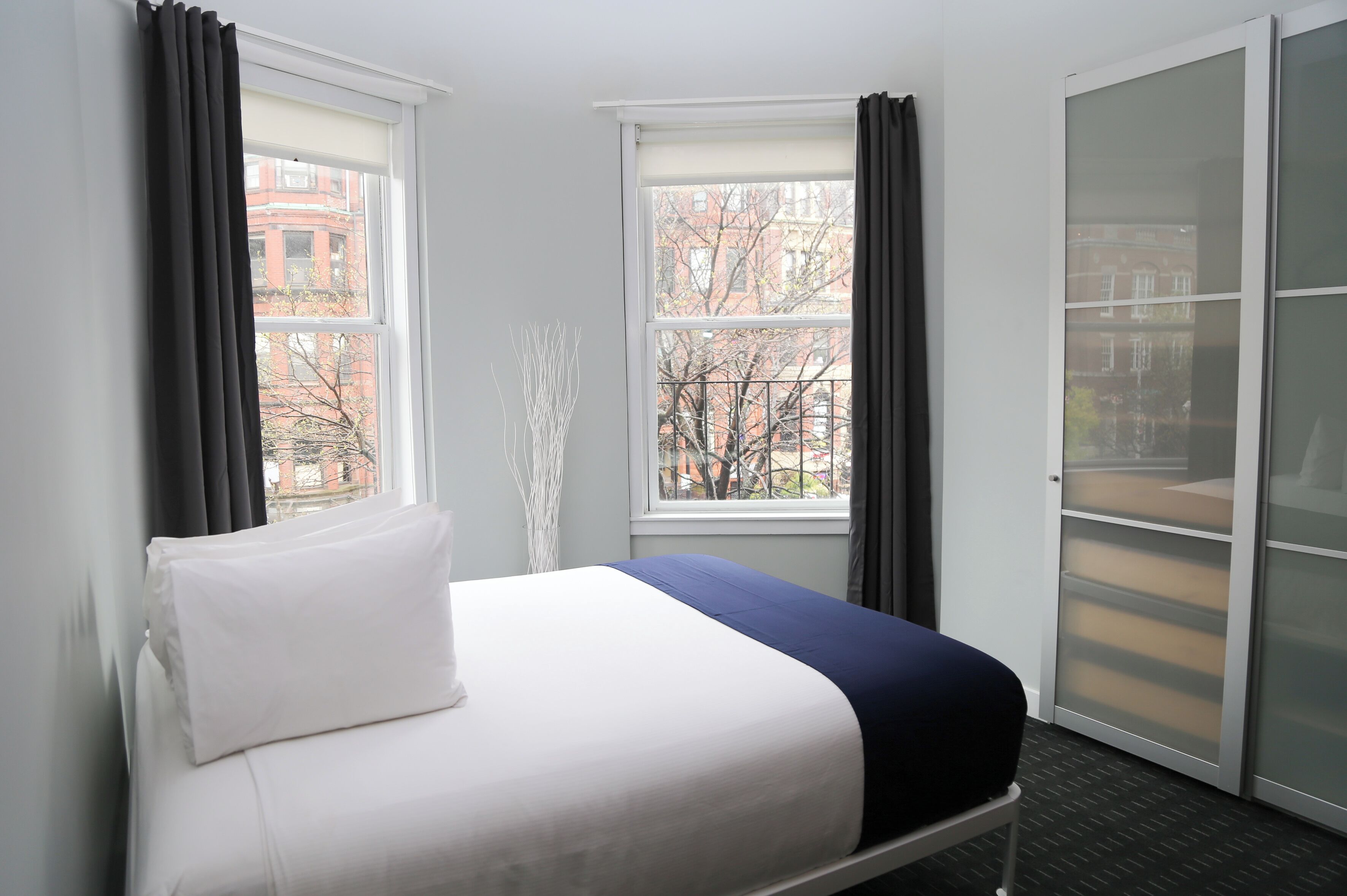 A hotel room with a bed, two windows, and a view of buildings outside.