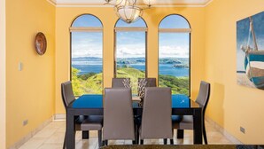 Een eetgedeelte met een tafel en stoelen, grote ramen met uitzicht op zee, en een kroonluchter.