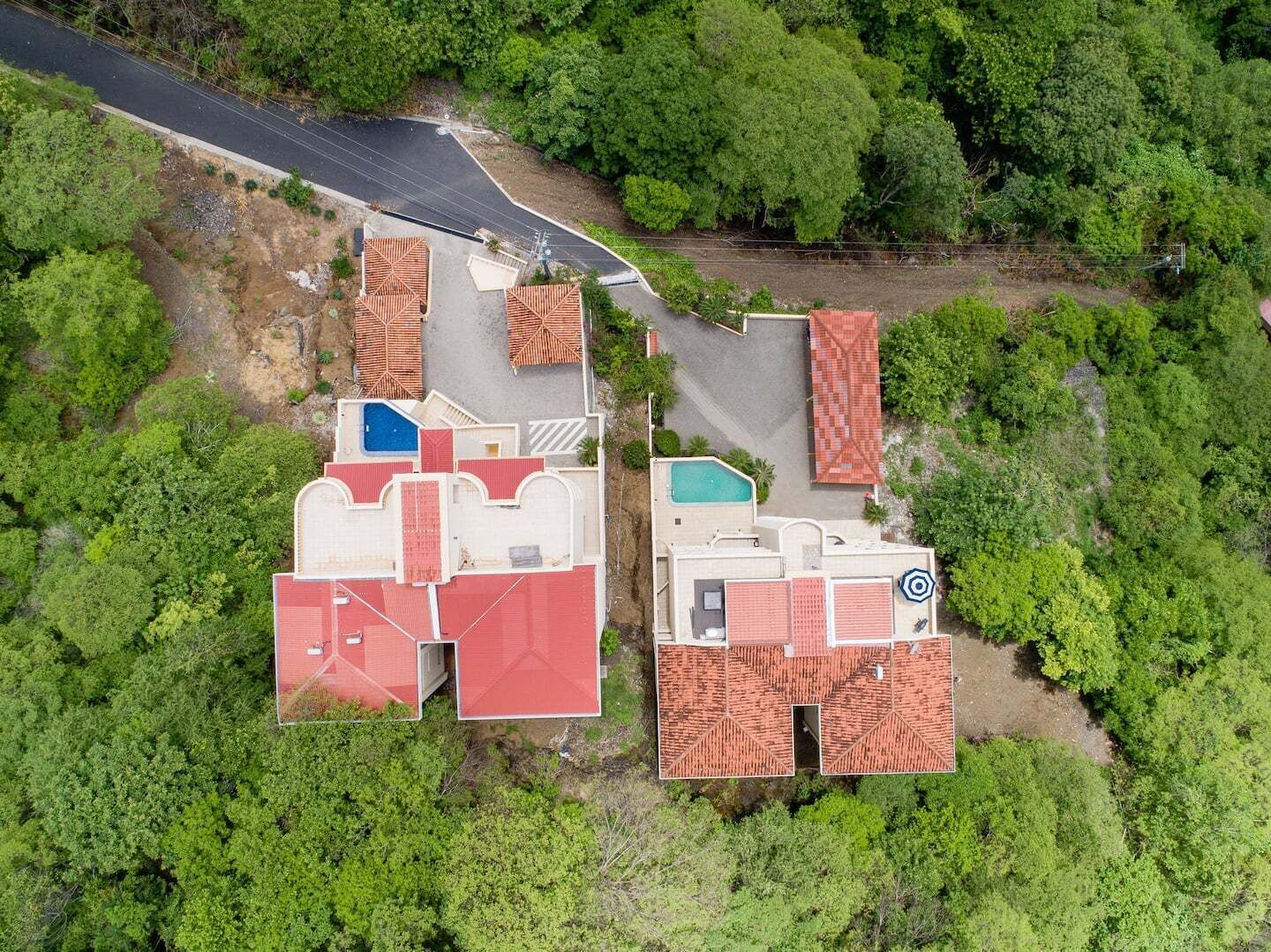 An aerial view of a property with multiple buildings, including a swimming pool and a tennis court, surrounded by dense greenery.