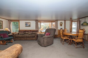 A cozy living room with a sectional sofa, a recliner, a dining table with chairs, and a view of autumn trees through large windows.