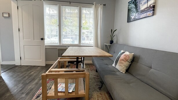 En stue med en grå sofa, et træsofabord, et træspisebord og et vindue med rullegardiner.