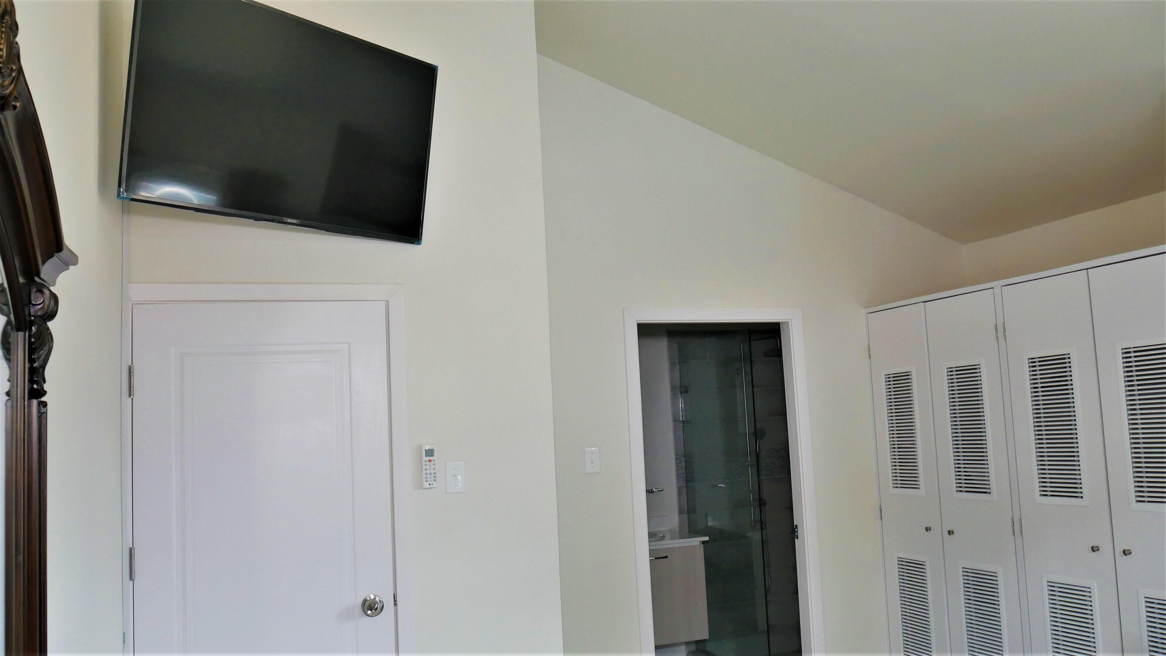 A room with a flat-screen TV mounted on the wall, a white door, a glass door leading to a bathroom, and a set of white lockers with ventilation slats.