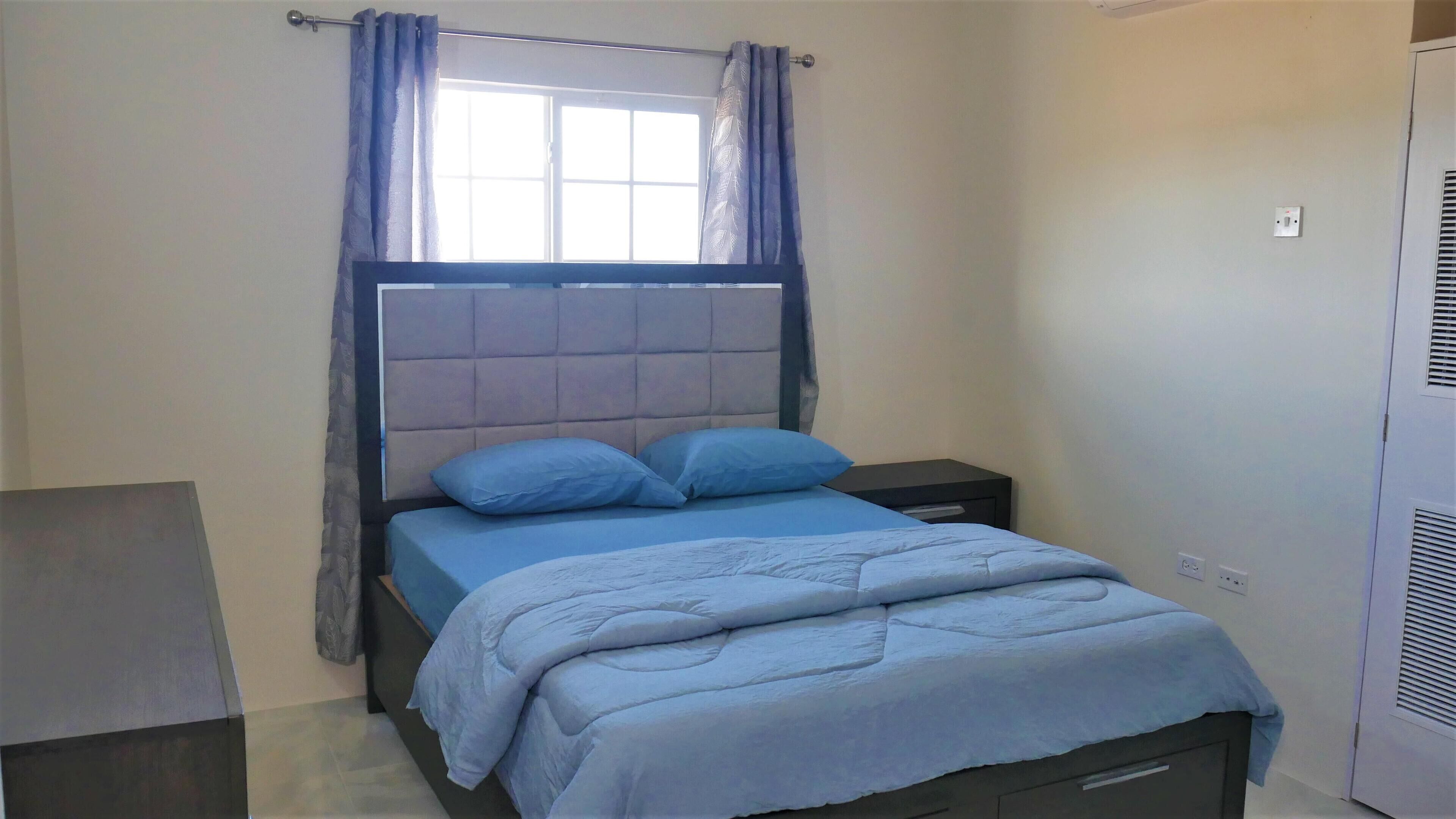 Un dormitorio con cama, mesita de noche, ventana con cortinas y aire acondicionado de pared.