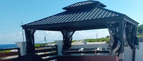 A covered outdoor seating area with a grill, benches, and a view of the ocean.