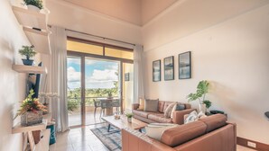A spacious living room with a brown leather sofa, a coffee table, and a dining area with a table and chairs. There are potted plants on shelves and a large window with curtains. - 1BR 4 Guests Golf View Course Casa de Campo (La Romana)