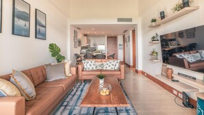 A spacious living room with a brown leather sofa, a wooden coffee table, and a blue patterned rug. A kitchen is visible in the background, and there are framed pictures on the walls. - 1BR 4 Guests Golf View Course Casa de Campo (La Romana)