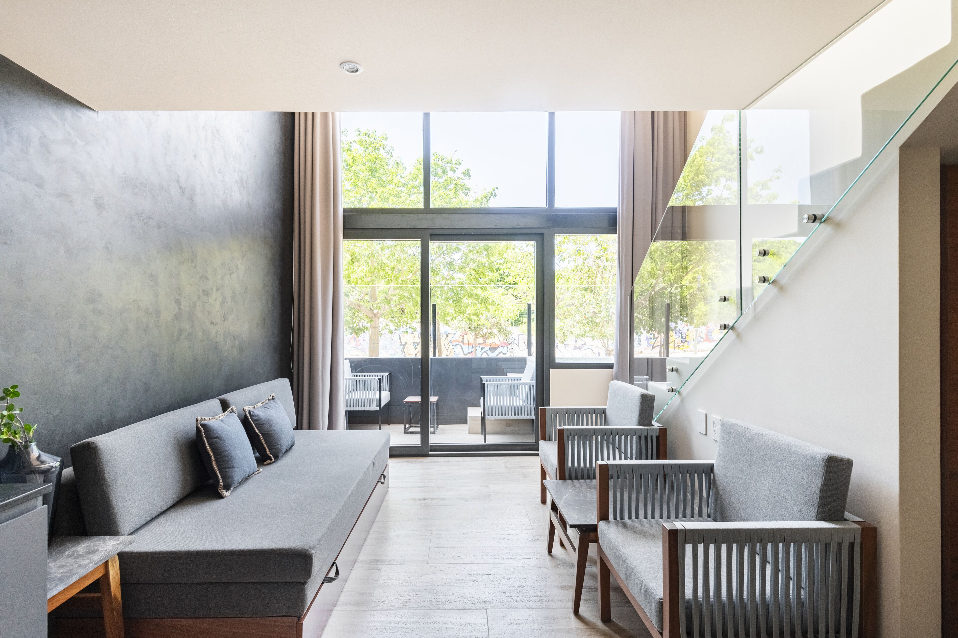 A modern interior with a grey sofa, wooden chairs, and a large window with a view of greenery.
